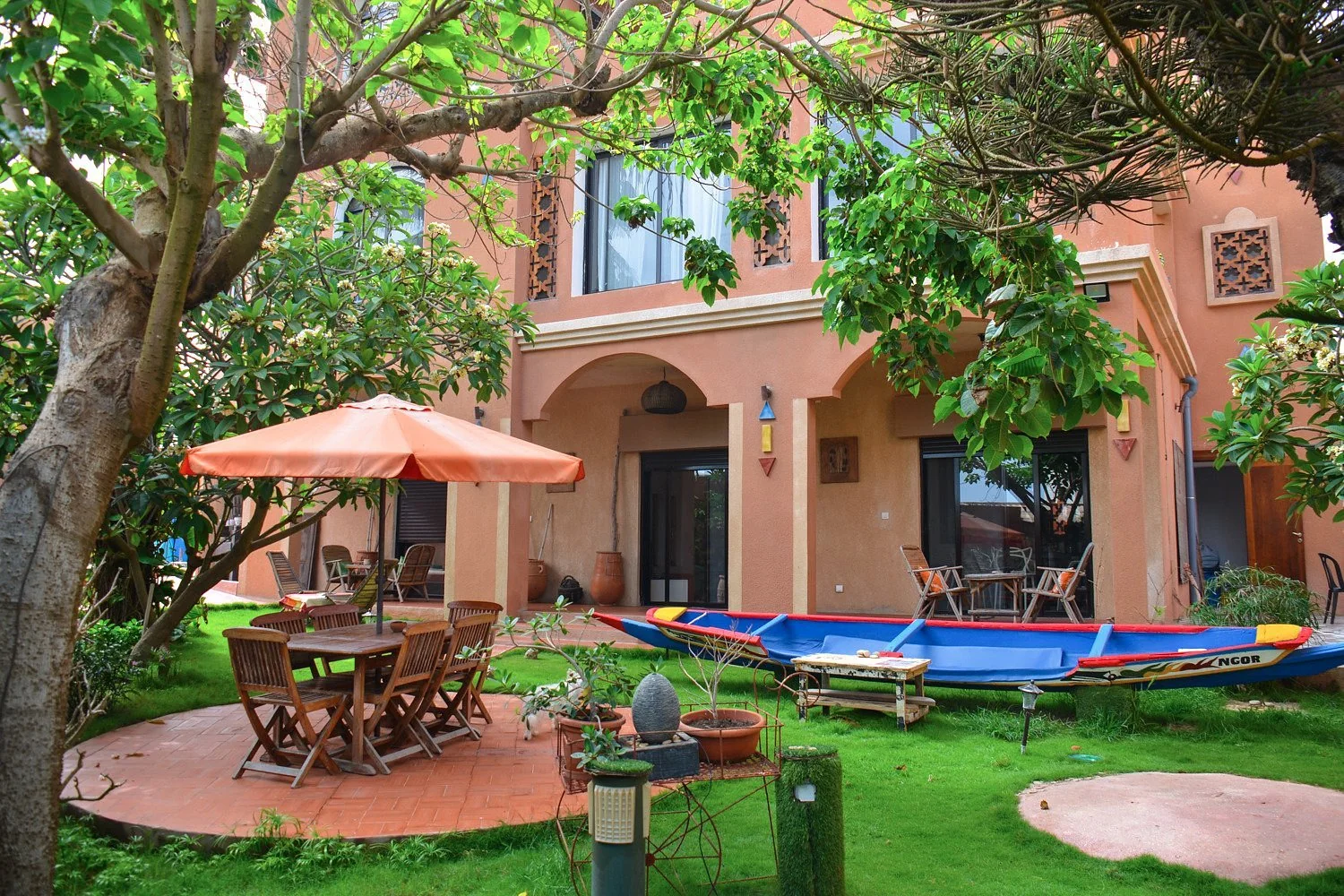 Garden lawn with a blue painted pirogue, shaded dining table and terracotta guest-house facade